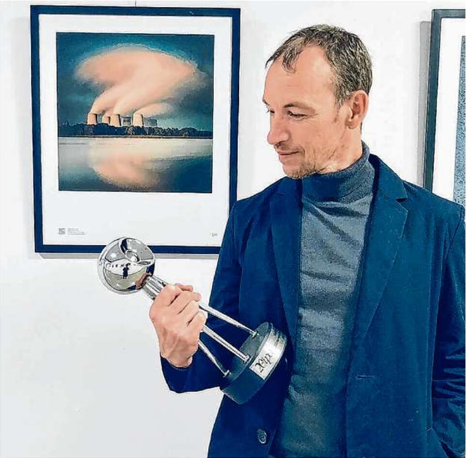 Steffen Reichardt hatte das Fotografieren schon aufgegeben und holt nun den globalen Foto-Award nach Cottbus. Hinter ihm ist sein Siegerfoto vom Kraftwerk Jänschwalde zu sehen.