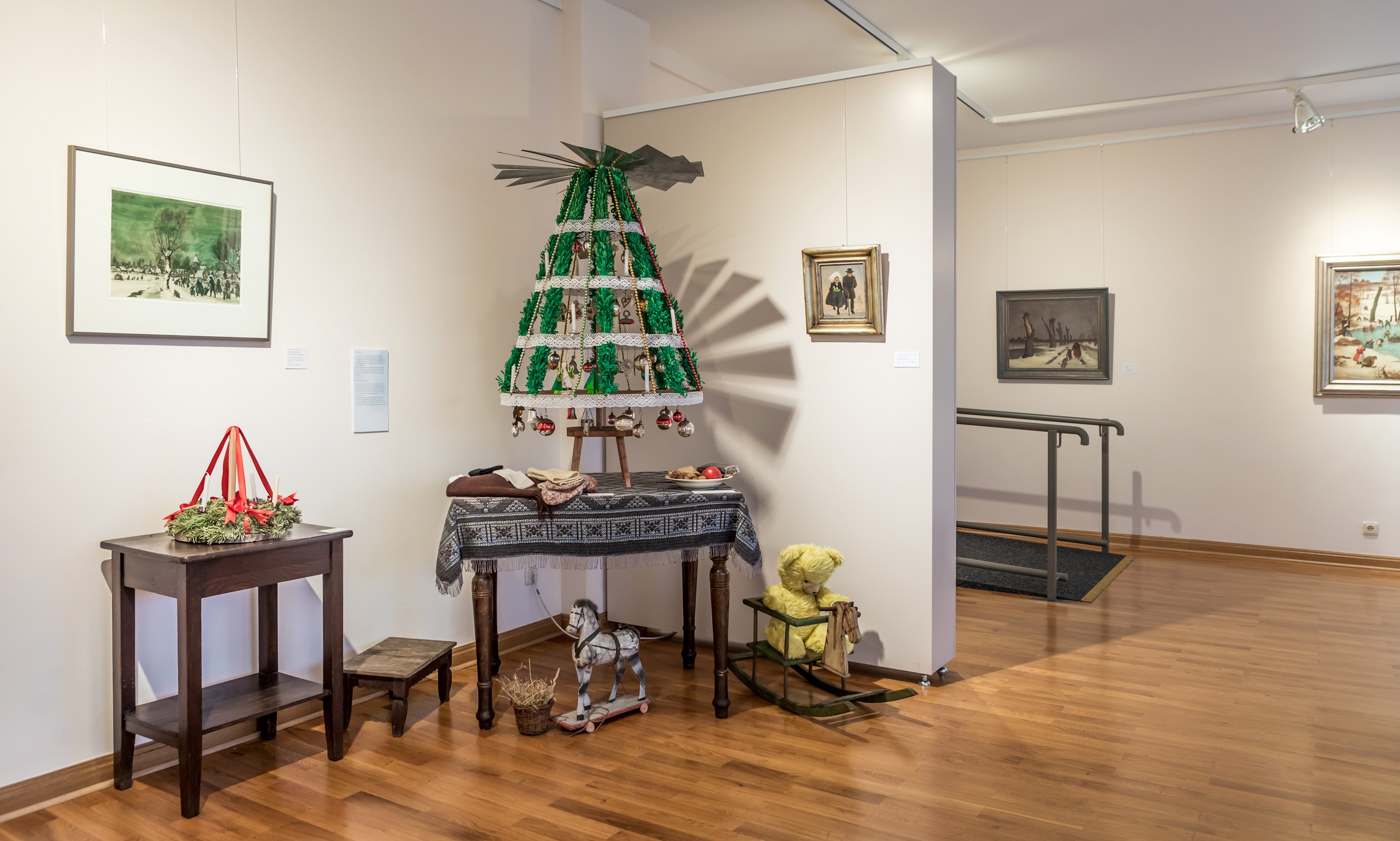 Drehbaum mit grünen und weißen Stoffbahnen auf einem Tisch, umgeben von Holzspielzeug und einem kleinen Tisch mit Kranz und roten Bändern in einem Ausstellungsraum mit Bildern an den Wänden.