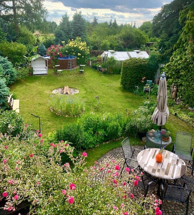 Blick aus der Vogelperspektive auf ein harmonisch angelegtes Gartenparadies mit blühenden Blumen, gepflegten Hecken, Bäumen und Sträuchern. Gewächshaus, gemütliche Sitzplätze, Feuerstelle und Planschpool laden zum Verweilen, Entspannen und Genießen ein.