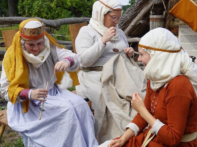 Drei Frauen in mittelalterlicher Kleidung sitzen im Freilichtmuseum. Sie nähen Kleidung mit der Hand und spinnen Fäden mit einer Spindel.